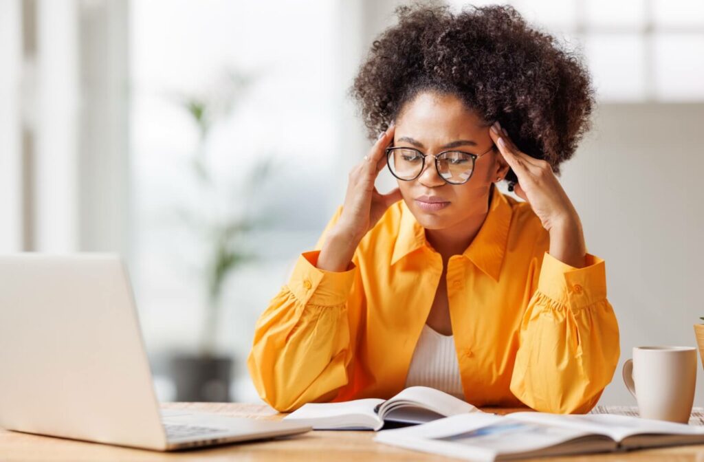 A young office worker with eyeglasses massages their temples while grimacing in pain from a headache